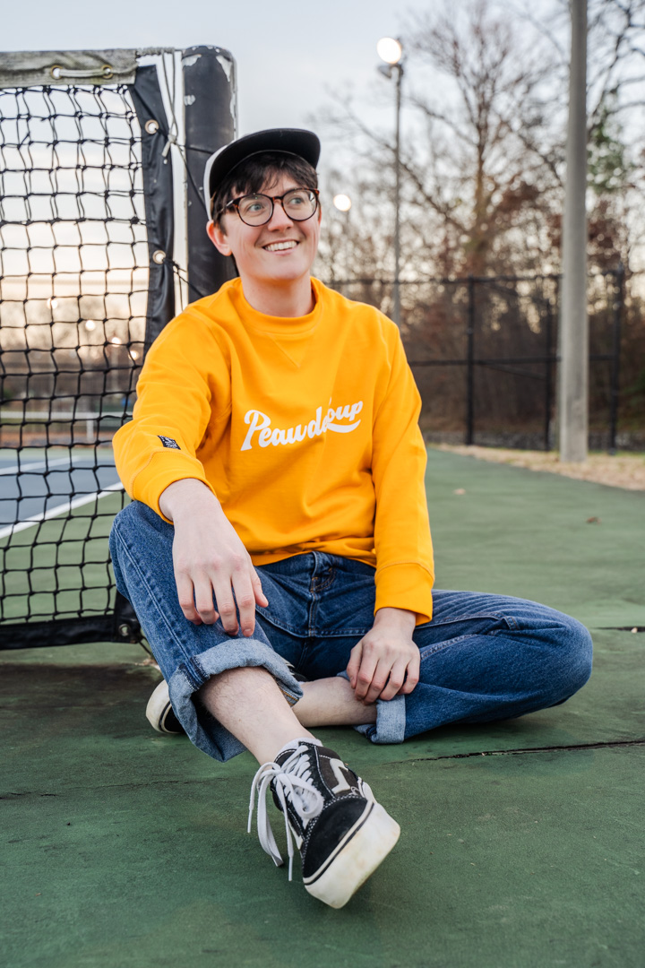 A young man wearing a yellow crewneck sweater, hat, and jeans sits smiling.